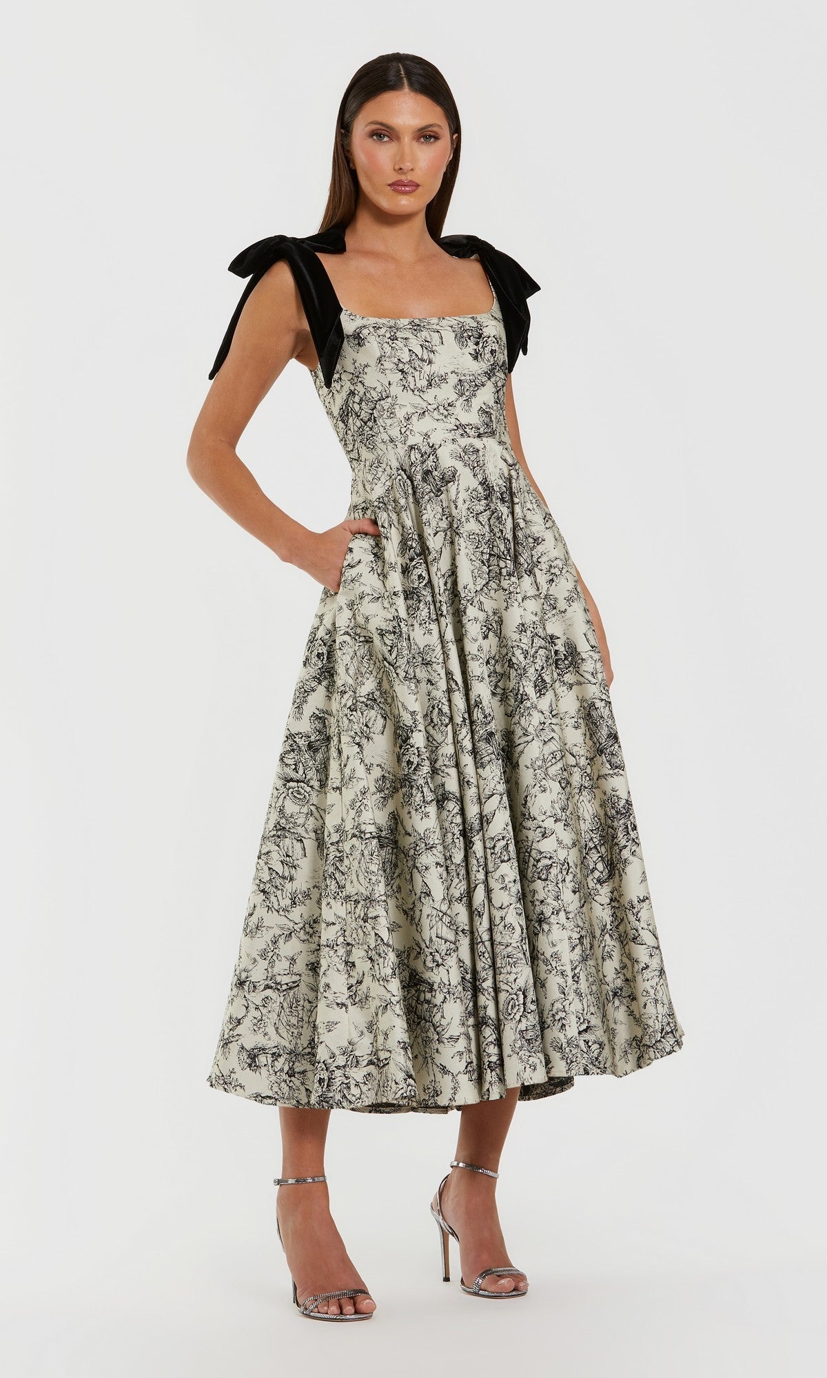 A woman wearing the Mac Duggal 27749 Midi Party Dress in Black Multi, featuring a sleeveless black-and-white floral print design with black bow shoulder straps and paired with metallic high-heeled sandals, stands against a plain white background.